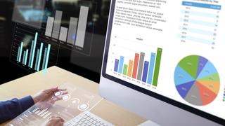 Business man or analyst in front of desktop computer holding a modern transparent tablet screen reviewing business performance and a return on investment, ROI, and investement risk analysis istock photo for BL